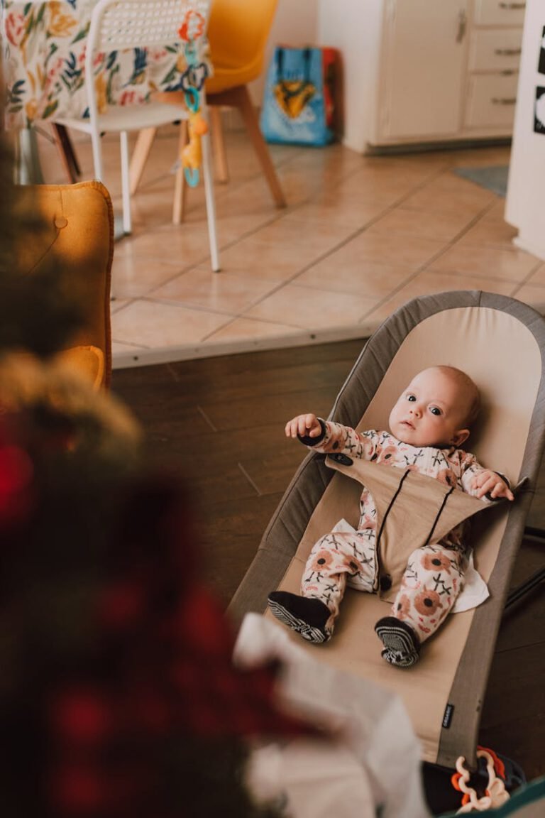 An infant sitting in a baby bouncer in the living room, looking at a blurred foreground object, most likely a Christmas tree.