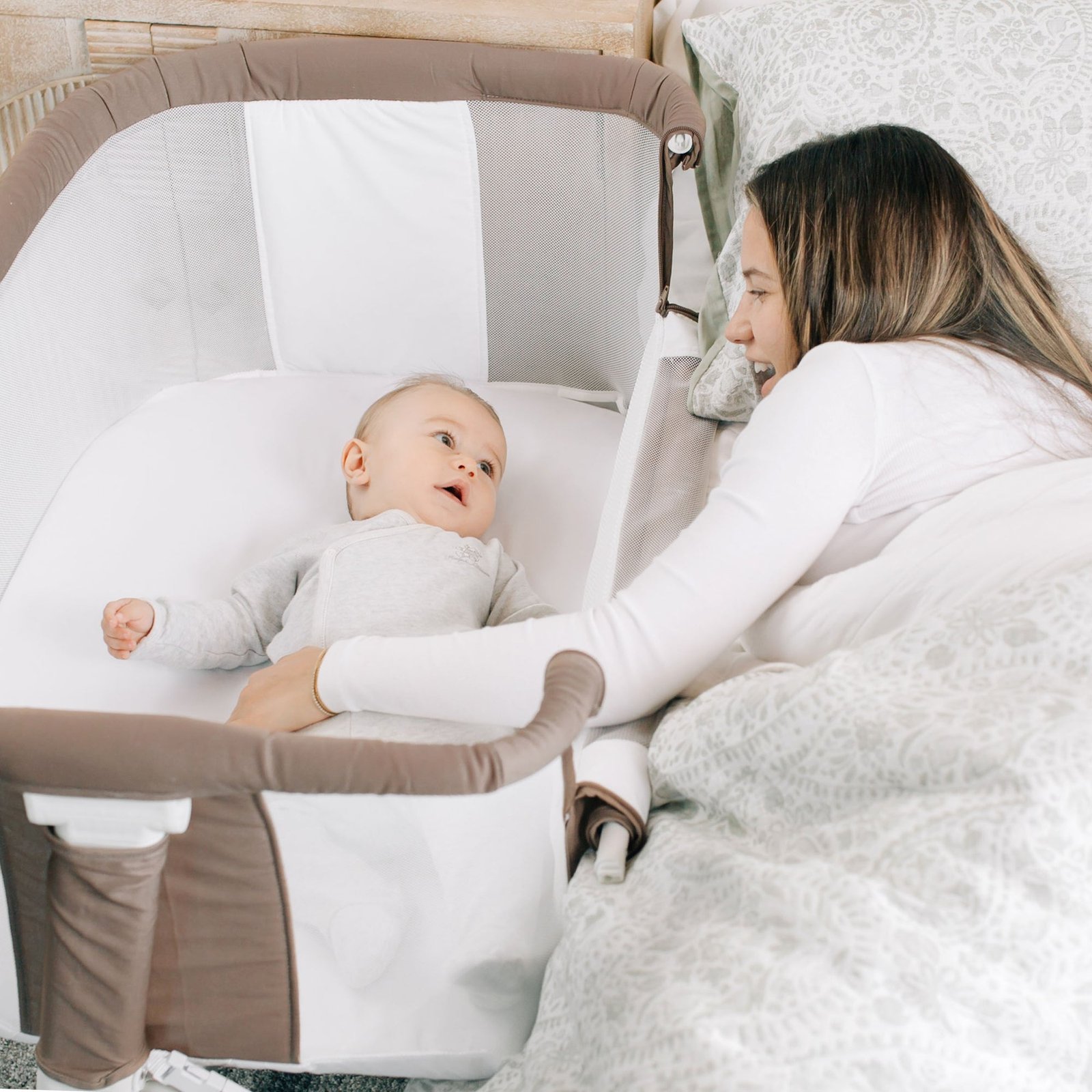 A mother lays in bed and speaks to her baby laying in the bedside bassinet.