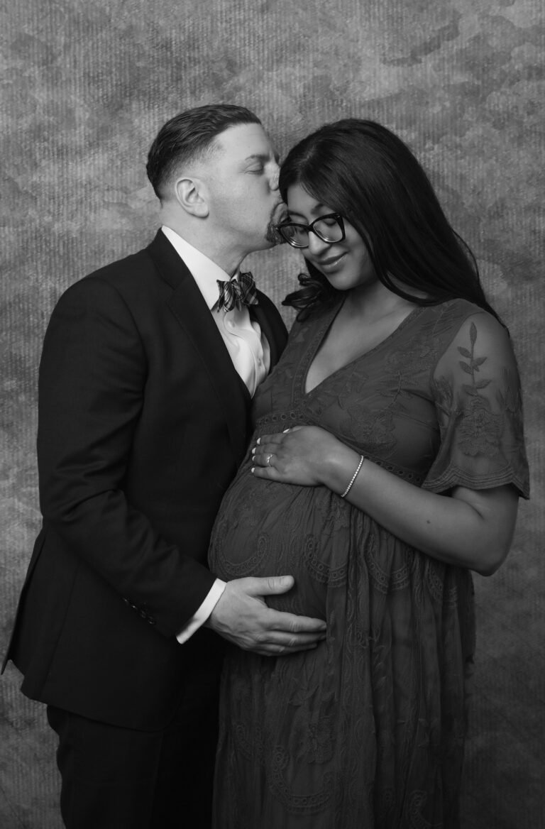 A black and white studio portrait of husband gently kissing his pregnant wife on the forehead while their hold her stomach together.